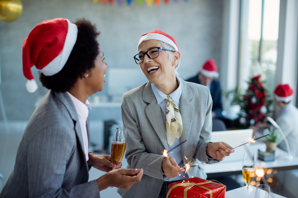 Two businesswomen in Christmas hats holding lit candles, smiling and celebrating the festive season together.