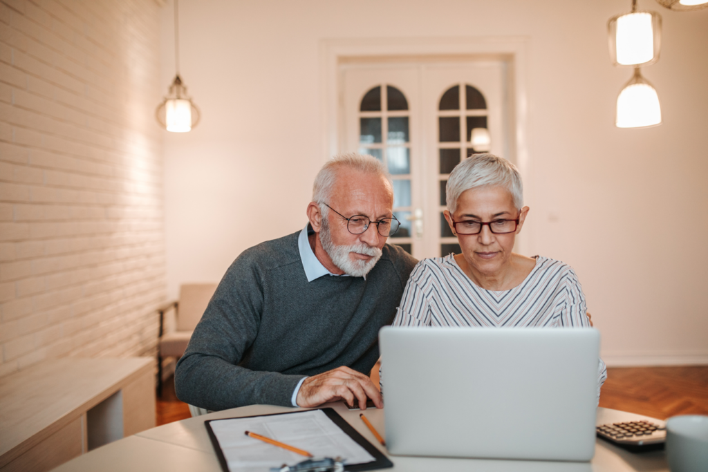 An elderly couple sitting together, smiling, and looking at a laptop screen.